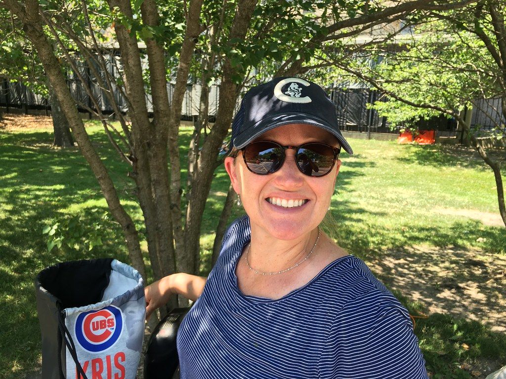 Smiling woman in a cap and sunglasses standing outdoors under shade trees