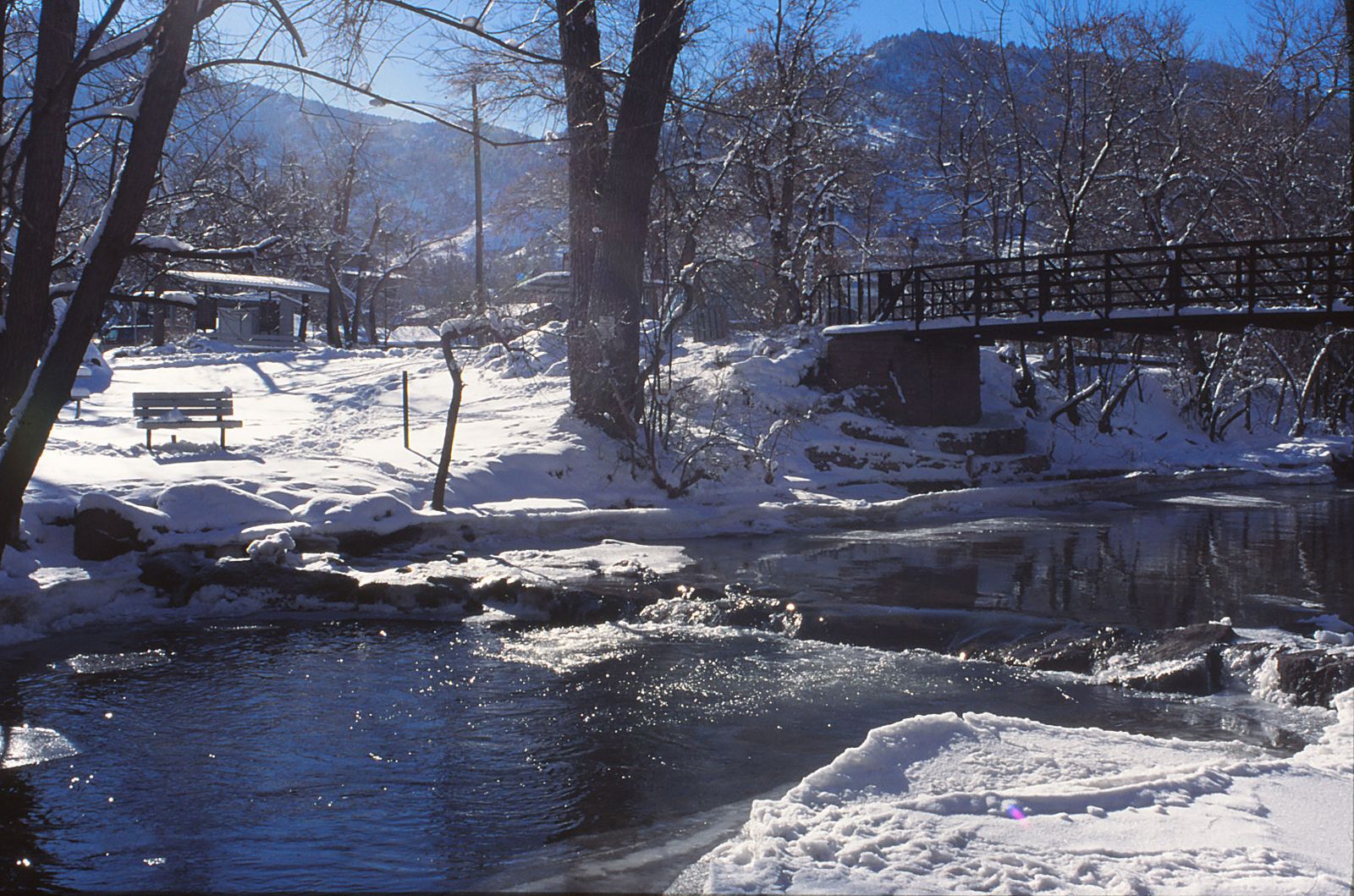 Creek running between snowy banks under bare trees with a footbridge in Boulder