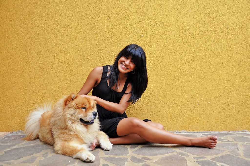 Smiling woman sitting on the ground beside her fluffy chow chow dog against a yellow wall