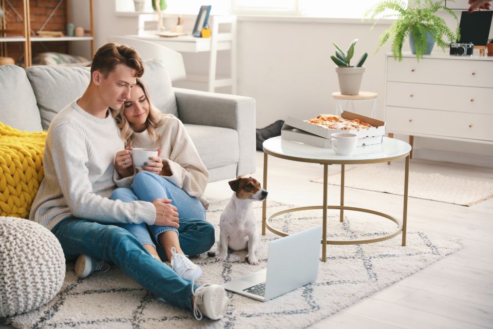 Couple sitting on a living room rug with a small dog and an open laptop