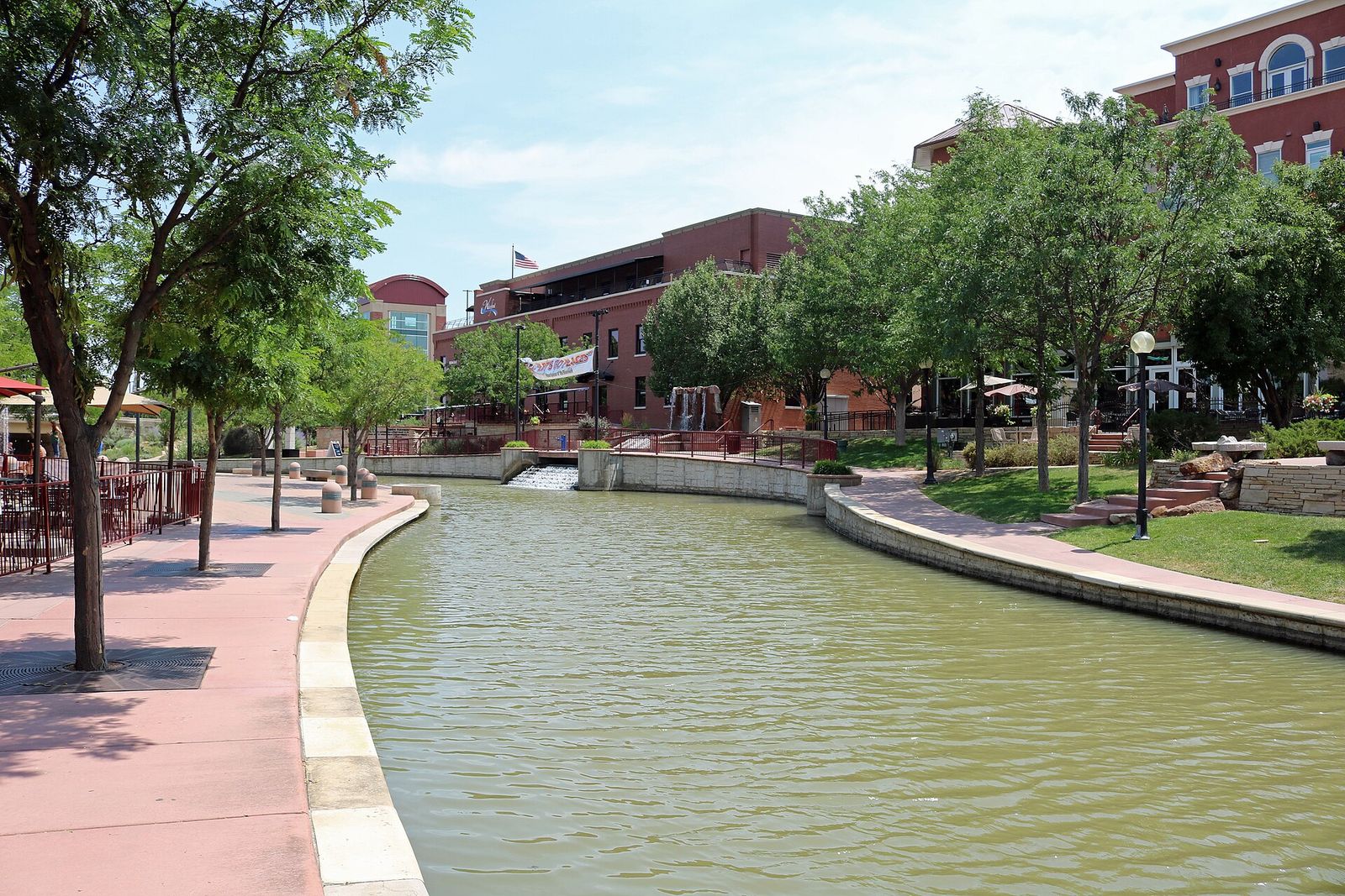 Channel of the Historic Arkansas Riverwalk of Pueblo with paved paths, trees and brick buildings