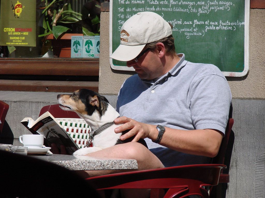 Man in a cap reading at a cafe table with a small dog resting in his lap