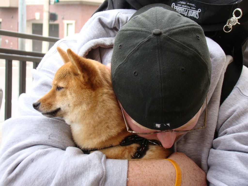 Man in a cap resting his head against a Shiba Inu in a warm hug