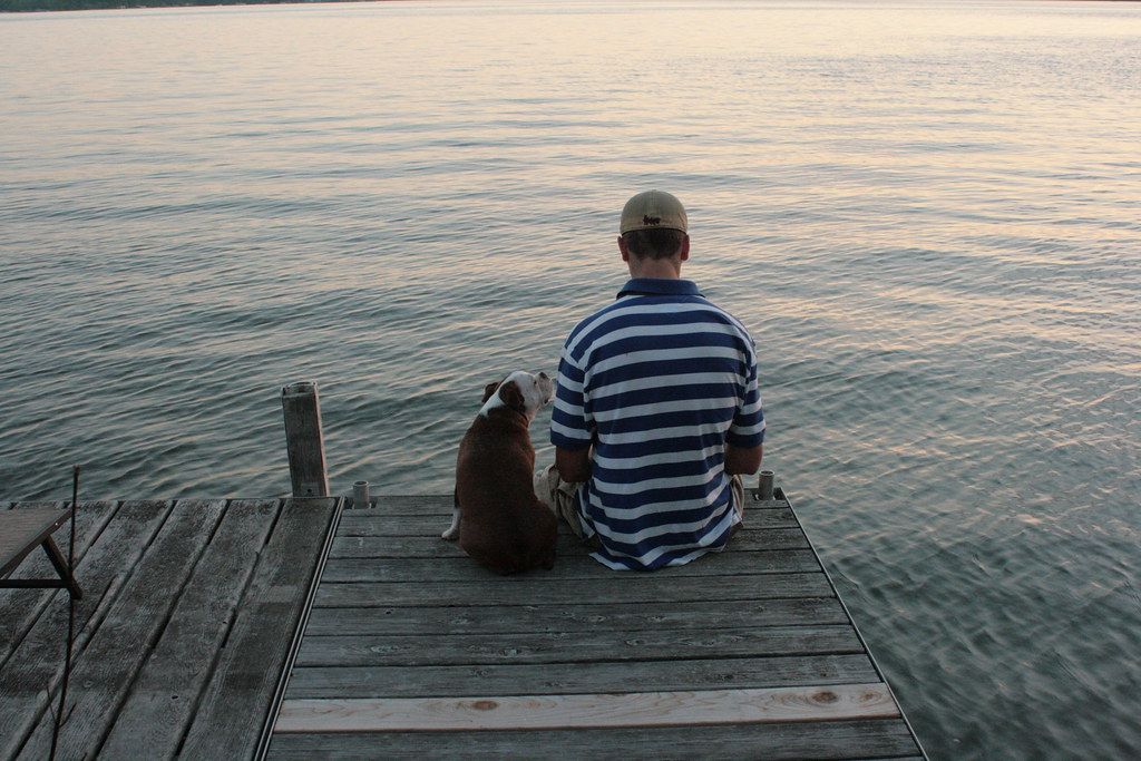 Man sitting on the end of a wooden dock beside his dog, looking out over calm water