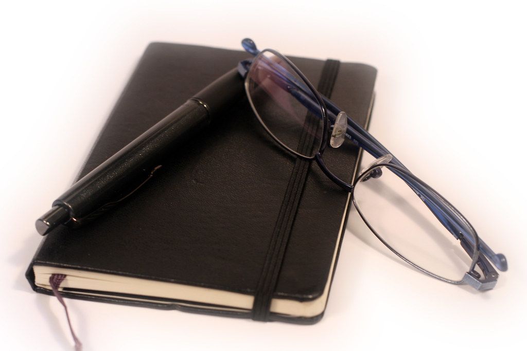 Black notebook with a pen and reading glasses on a white surface