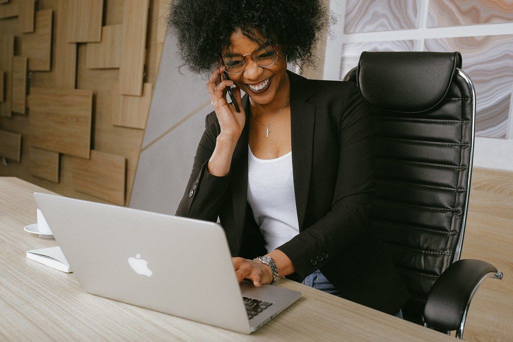 Smiling woman in a blazer talking on the phone at a desk with a laptop