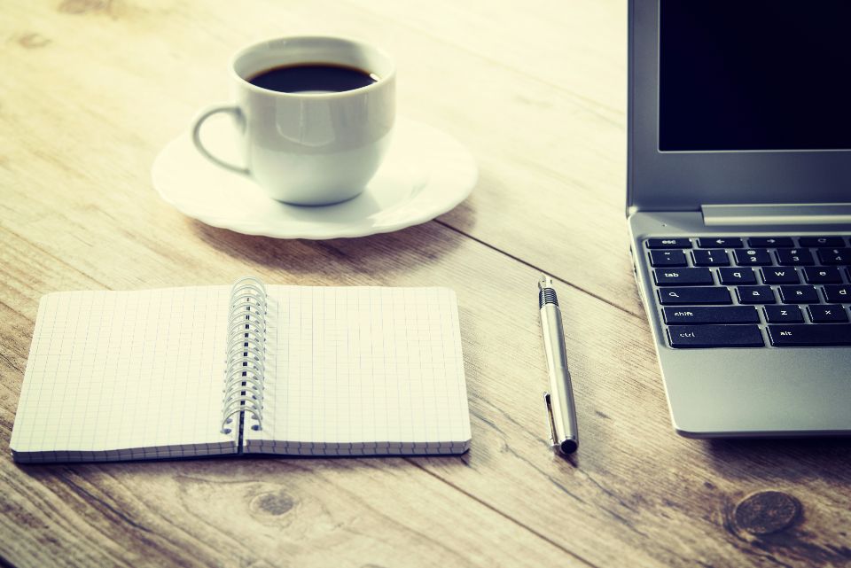 Laptop, open spiral notebook, pen and a cup of coffee on a wooden desk