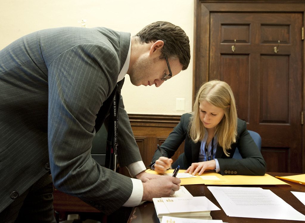 Woman signing papers at a wooden desk while a man leans in to point at the page