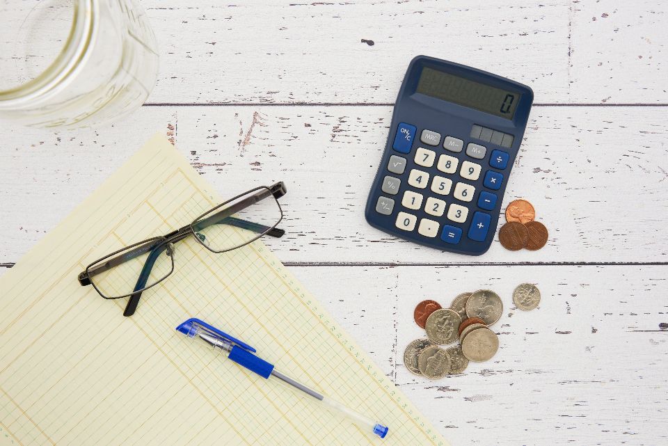 Blue calculator, eyeglasses, a pen and loose coins on a white wooden desk