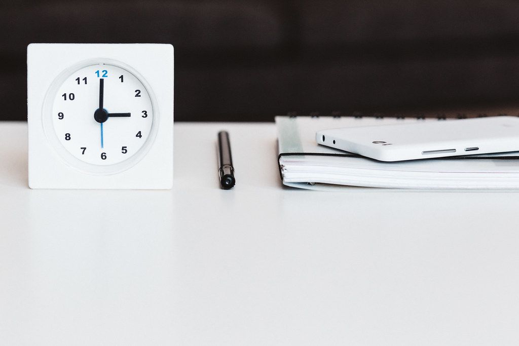 White desk clock beside a pen and a closed notebook on a white desk