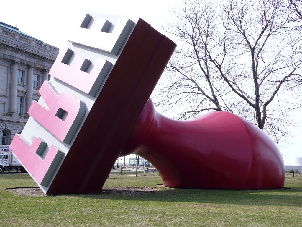 Oversized public sculpture of a red rubber stamp tipped over on a city lawn