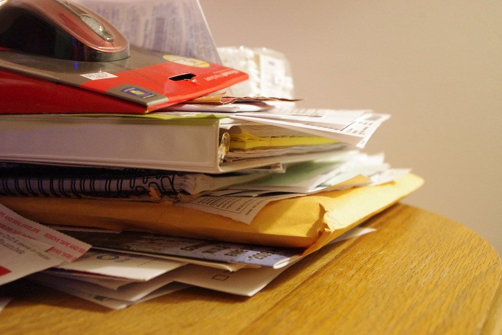 Messy stack of folders, envelopes and loose paperwork on a wooden desk