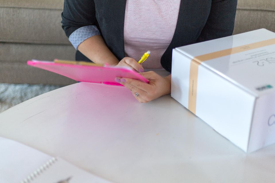 Hand writing notes on a pink clipboard next to a white box