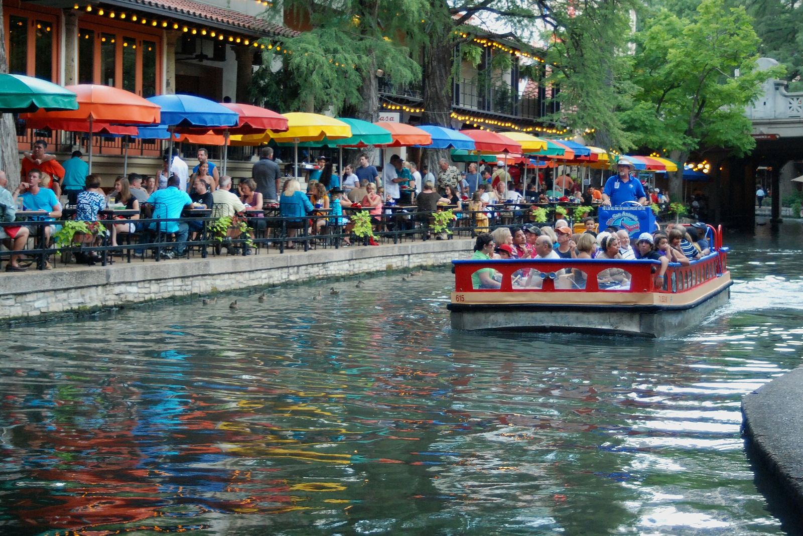 Tour boat passing restaurant patios with colorful umbrellas on the San Antonio River Walk